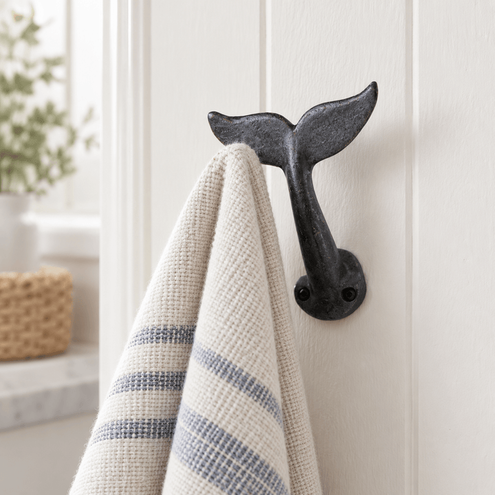 Black cast iron whale tail wall hook holding a striped hand towel in a bright coastal bathroom