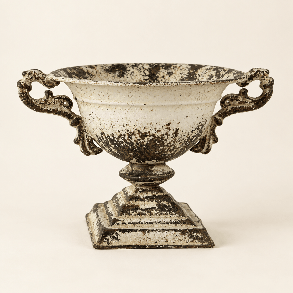 Distressed white iron pedestal urn with ornate handles and square base against an ivory backdrop