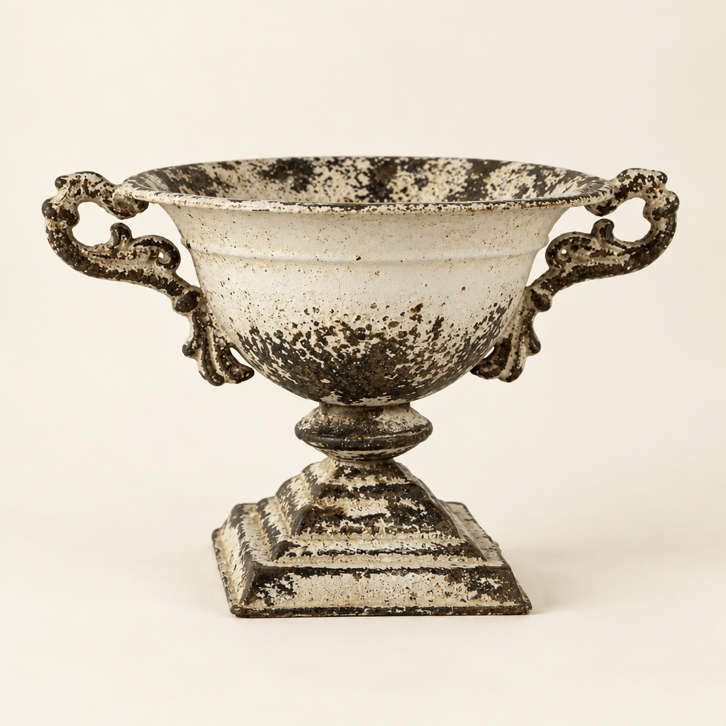 Distressed white iron pedestal urn with ornate handles and square base against an ivory backdrop