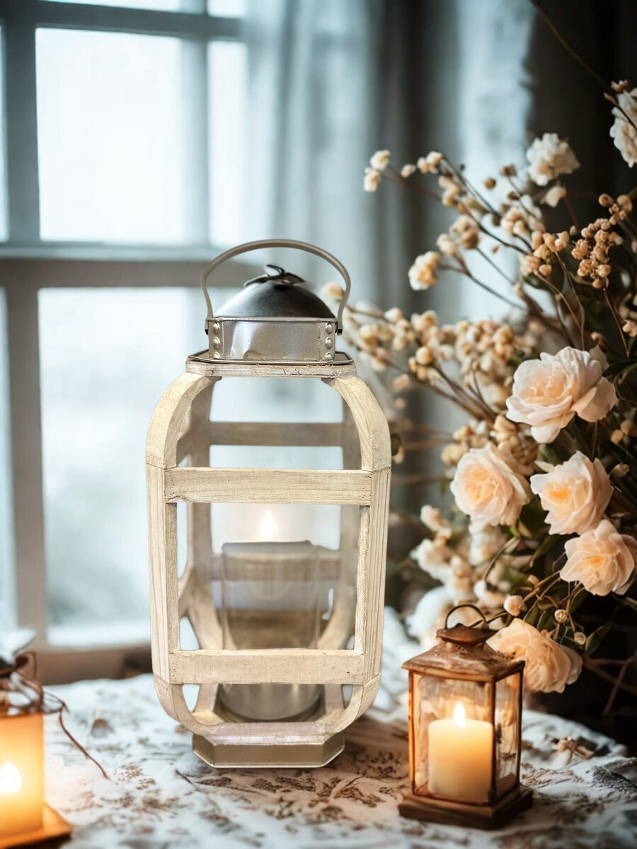 Distressed white wood and metal candle lantern glowing beside flowers in a window