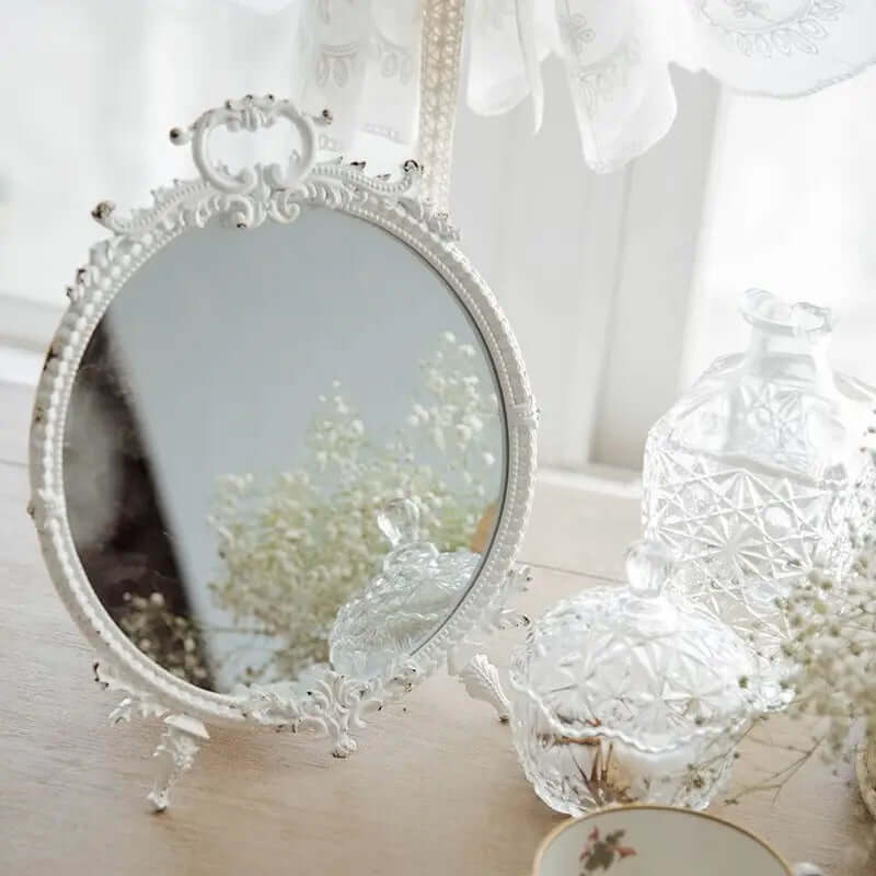 Vintage makeup mirror with distressed white finish and ornate pewter frame on a wooden vanity, reflecting elegant decor.