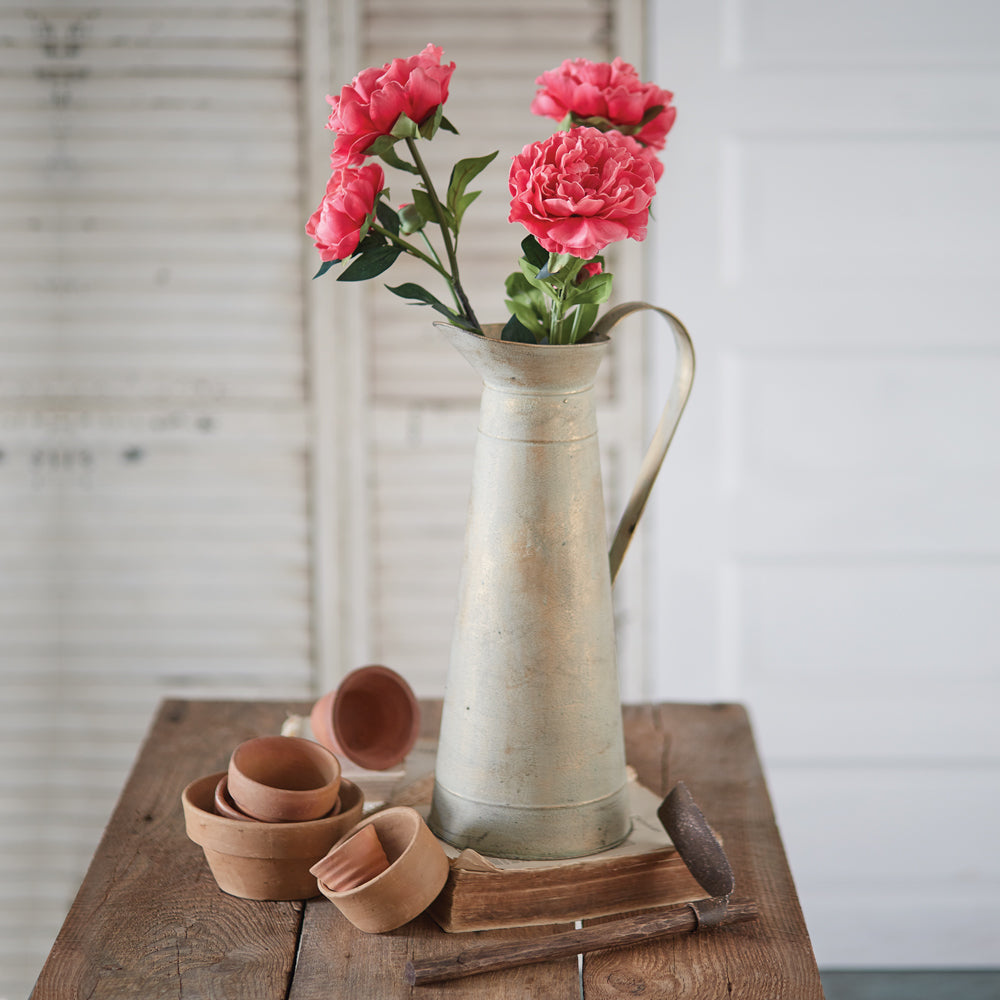 Faux pink peony flowers in a rustic vase, surrounded by terracotta pots on a wooden table for cottagecore decor.
