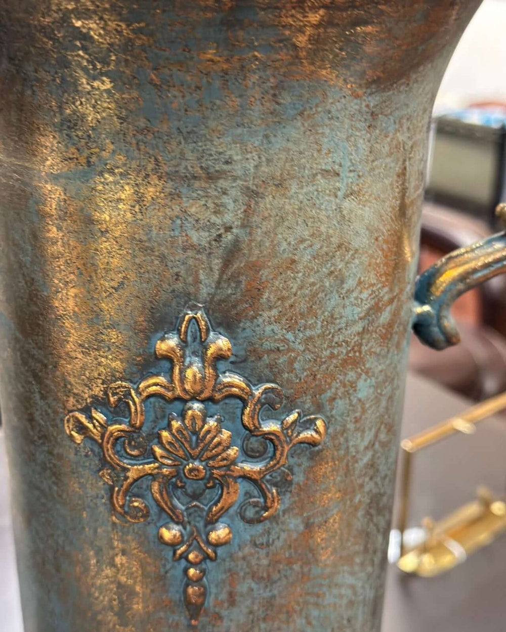 Ornate French country antique gold urn vase with an embossed floral medallion and curved handle, evoking classic elegance.
