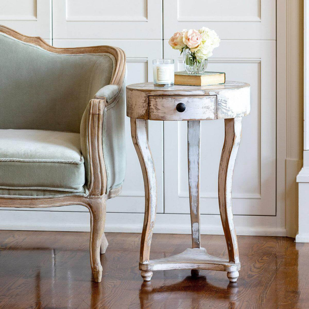 French Country Round Accent Table with Drawer in antique white wash next to an elegant armchair, showcasing provincial charm.