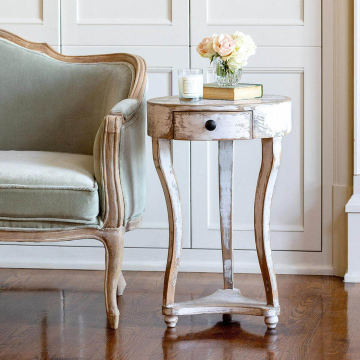 French Country Round Accent Table with Drawer in antique white wash next to an elegant armchair, showcasing provincial charm.