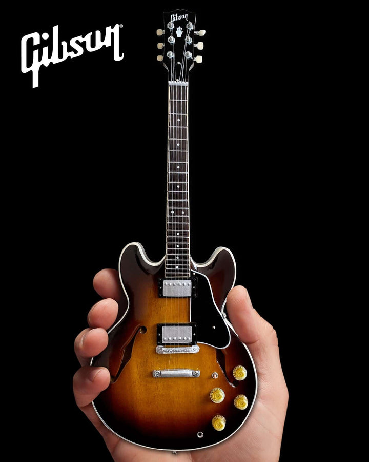 Hand holding a Gibson ES-335 Vintage Sunburst Mini Guitar Replica against a black background.
