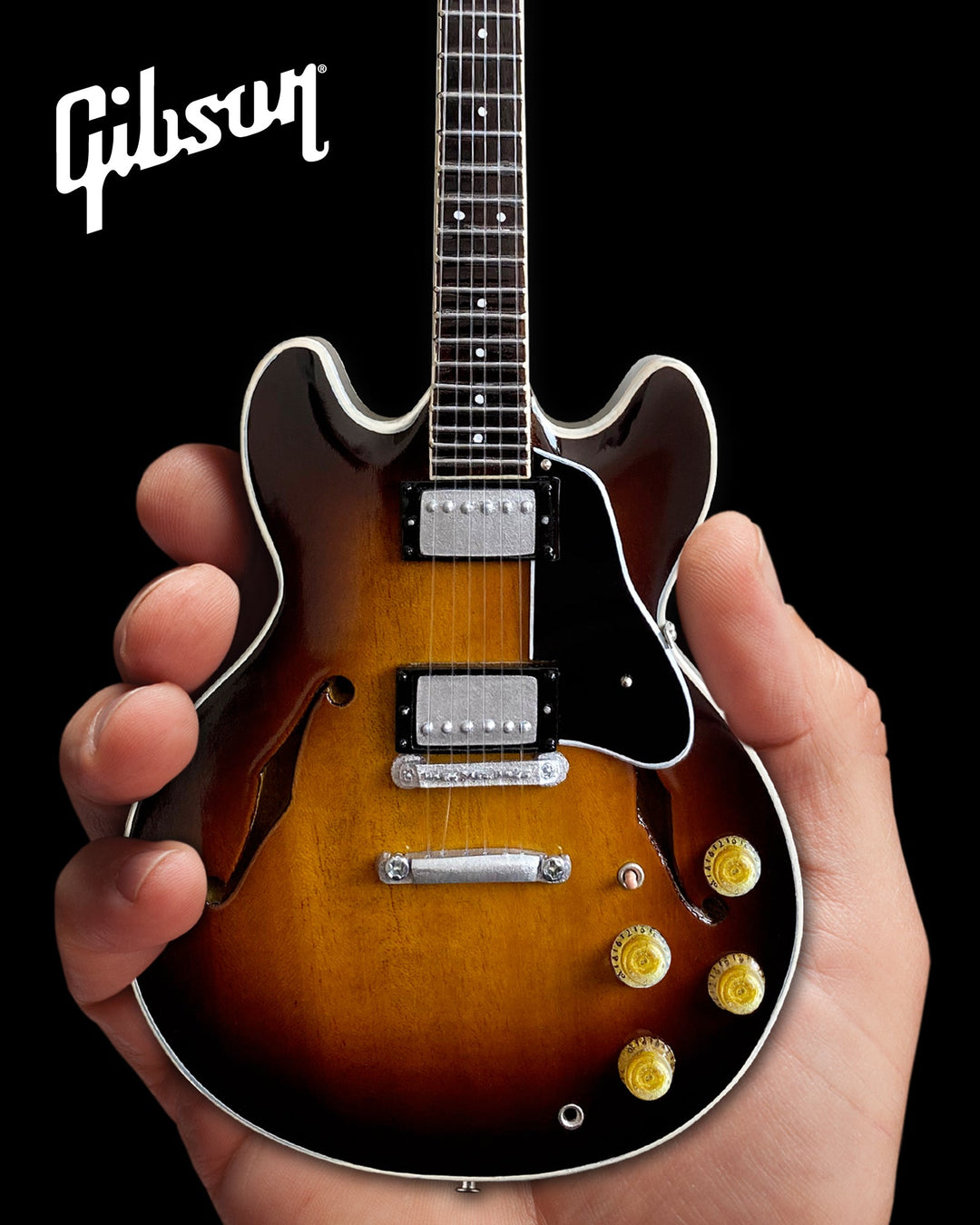 Hand holding a Gibson ES-335 Vintage Sunburst Mini Guitar Replica against a black background.