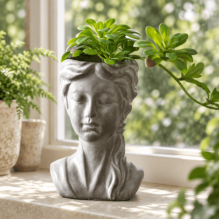 Gray cement girl bust planter with braided hair and succulent foliage on a pale stone windowsill.