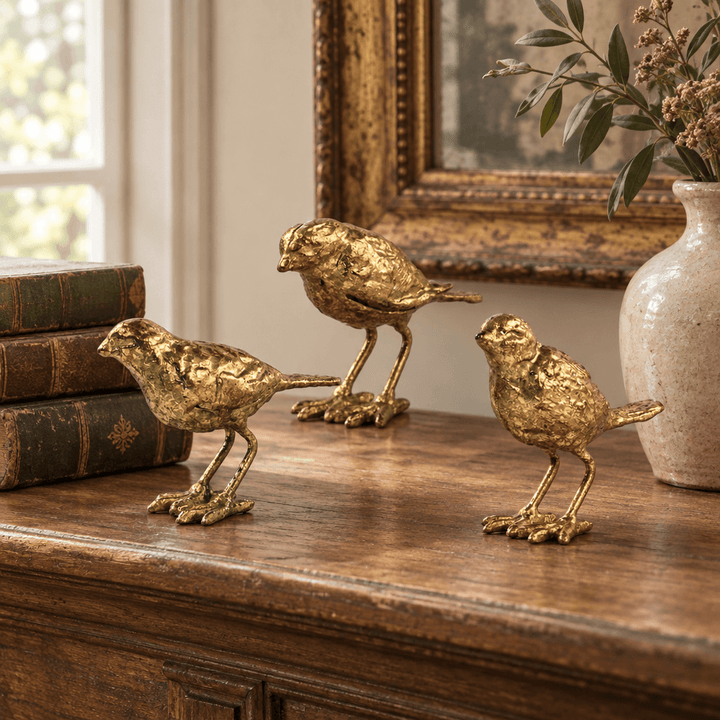 Three gold-leaf bird figurines styled on a wooden mantel beside books and a ceramic vase.