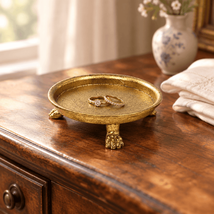 Gold-leaf pewter claw-foot dish styled with rings on a wooden dressing table beside ivory linen.