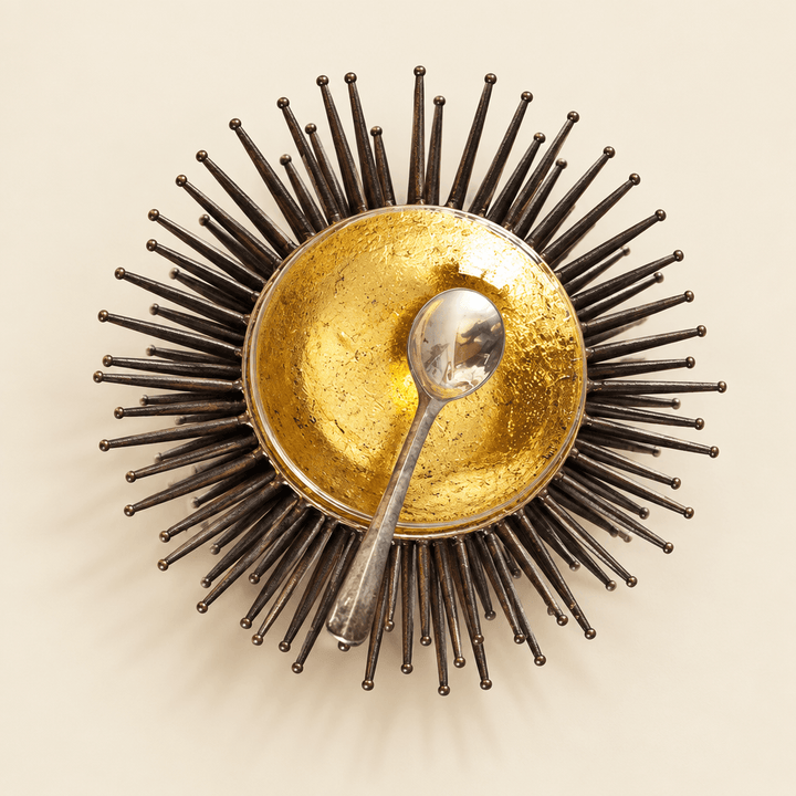 Gold leaf sea urchin accent bowl with dark iron spokes and silver spoon on an ivory studio background
