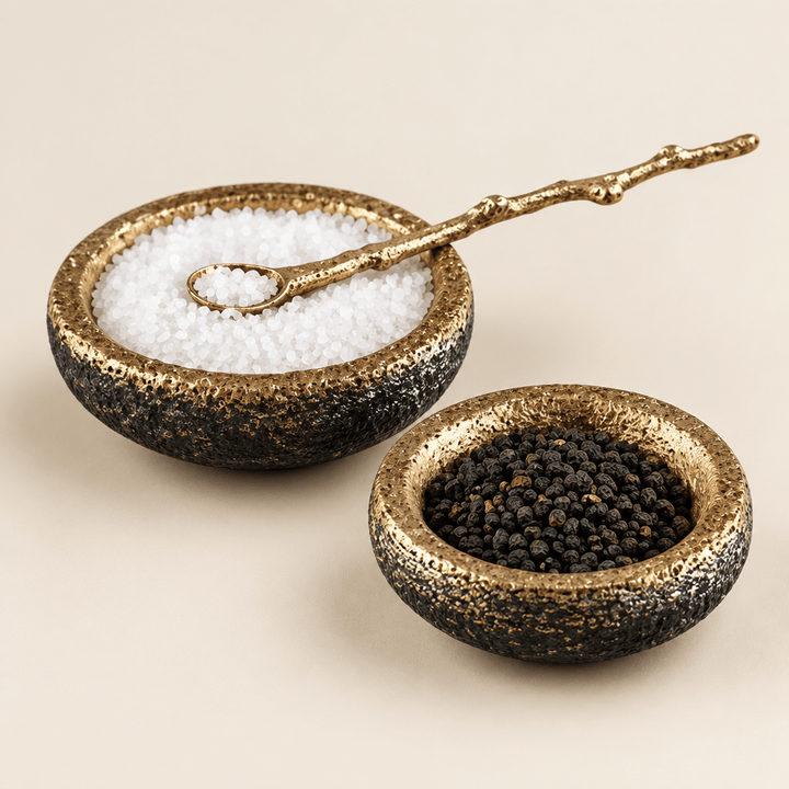 Two textured gold-toned salt and pepper cellars with one delicate gold spoon on an ivory background