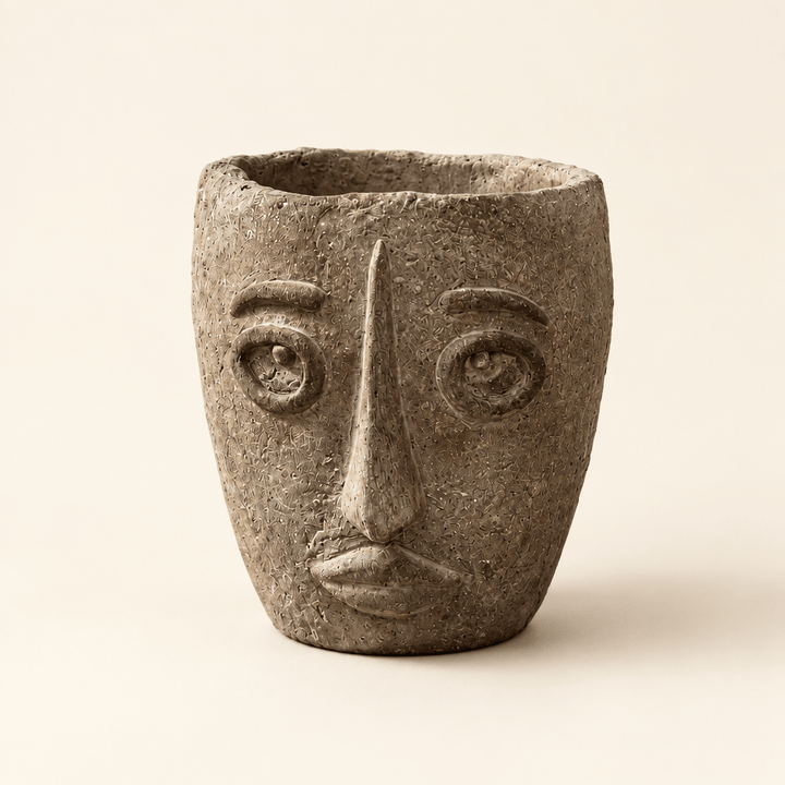 Earthy brown-gray cement face planter with raised eyes, a long nose, and sculpted lips on an ivory background
