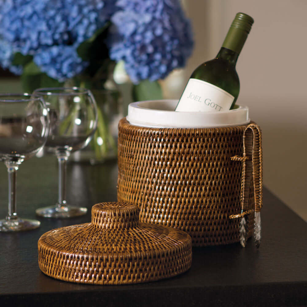 Handwoven vintage rattan ice bucket with lid and tongs, elegantly holding a wine bottle on a stylish table.