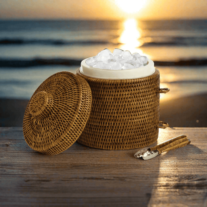 Handwoven vintage rattan ice bucket with lid and tongs filled with ice against a sunset backdrop.