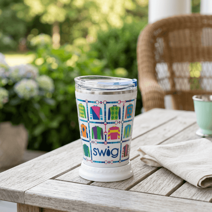 Colorful equestrian mint julep tumbler on a wooden porch table with cream linen and a garden backdrop.