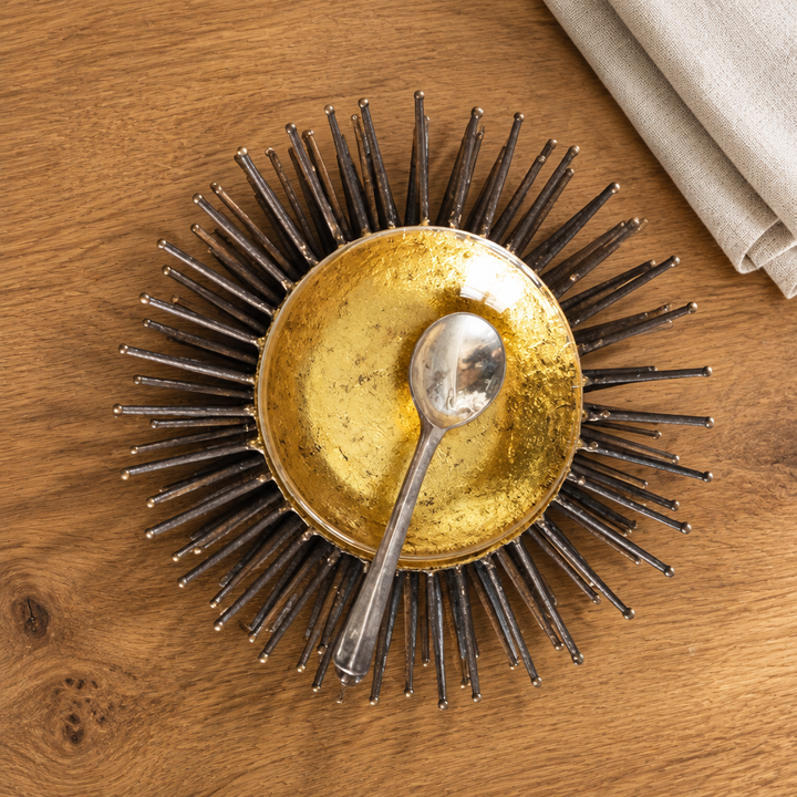 Overhead view of gold leaf sea urchin accent bowl and silver spoon on oak beside ivory linen