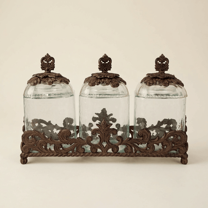 Three empty Bellavita glass canisters with ornate dark metal lids and shared scrollwork base on an ivory background