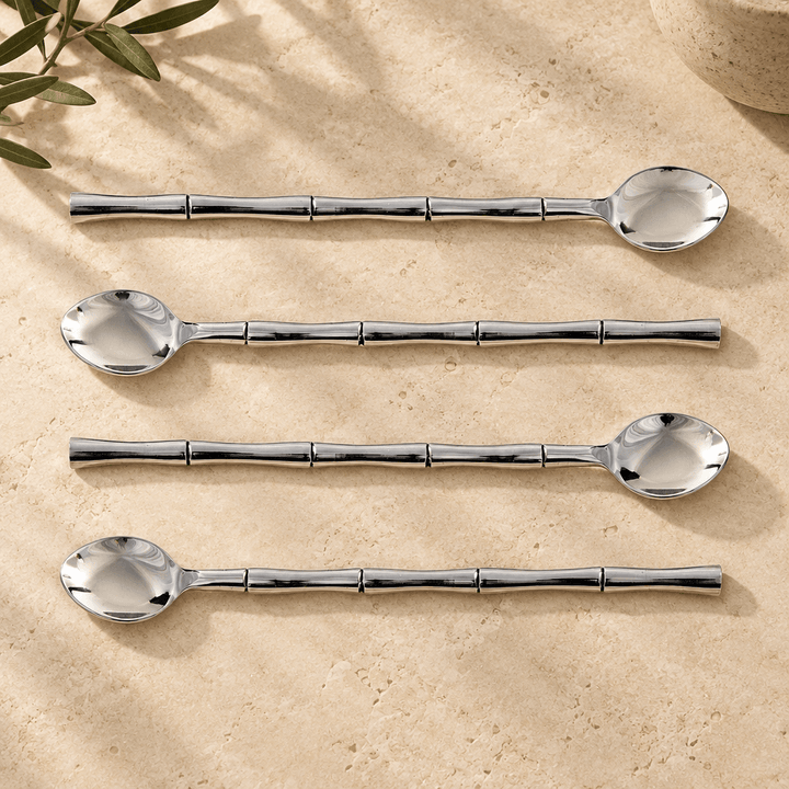 Four silver stainless steel stirring spoons with bamboo-style handles arranged on ivory limestone.