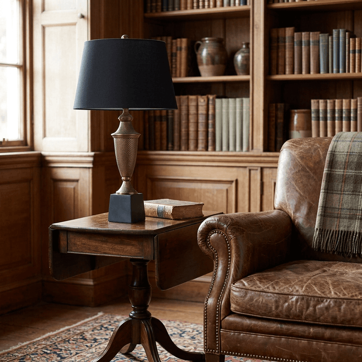 Ashbourne urn table lamp with textured antique brass finish and black shade beside a leather armchair in a library setting.