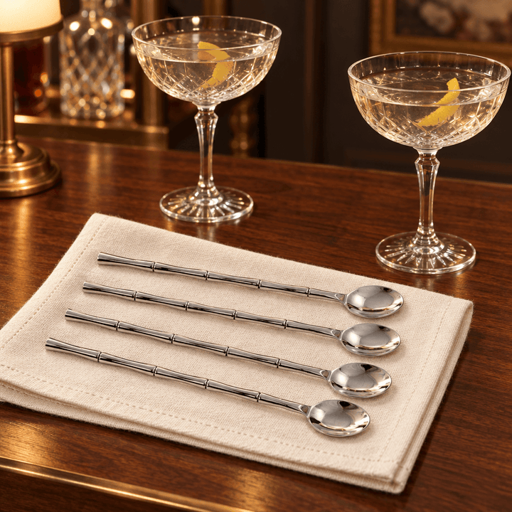 Bamboo-handle silver stirring spoons styled on cream linen beside cocktail glasses on a walnut home bar.
