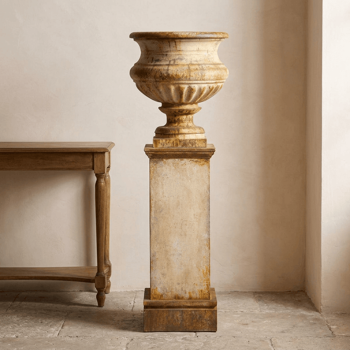 Montclair Iron Urn with Pedestal in Rust Patina, showing its pale aged finish and warm rust detailing beside a wooden console.