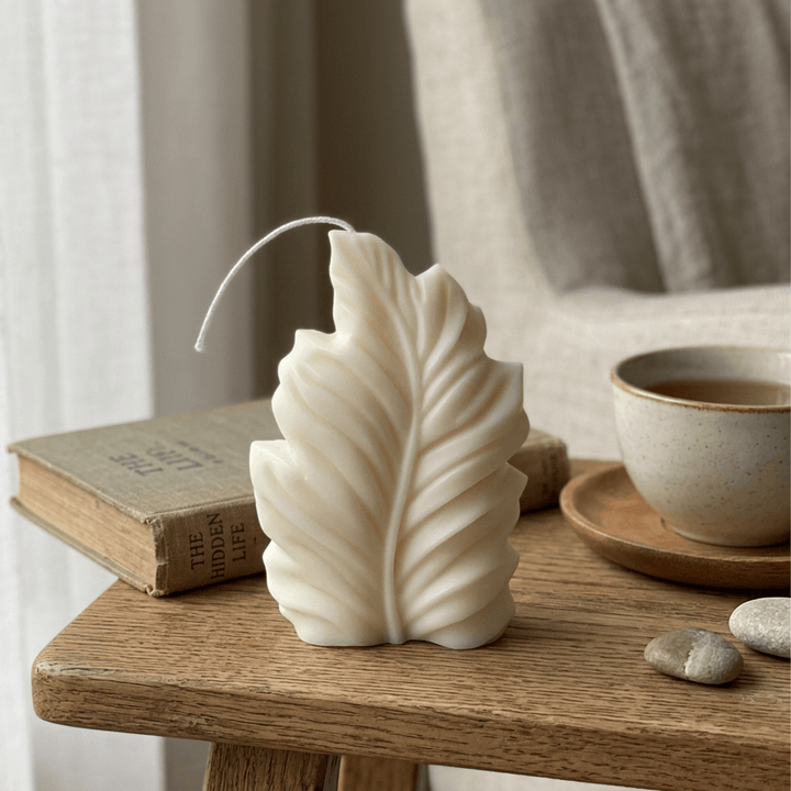 Ivory feather-shaped soy wax candle on an oak side table beside a book and tea cup in a reading nook.