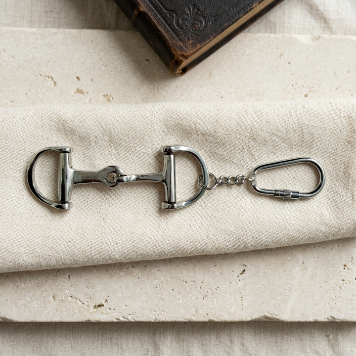 Silver D-ring snaffle bit keychain with a carabiner-style ring displayed on cream linen over pale stone.