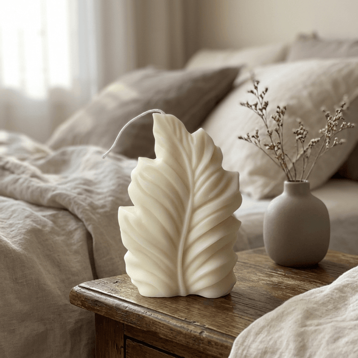 Ivory feather-shaped soy wax candle on a wood bedside table with linen bedding and a ceramic vase.