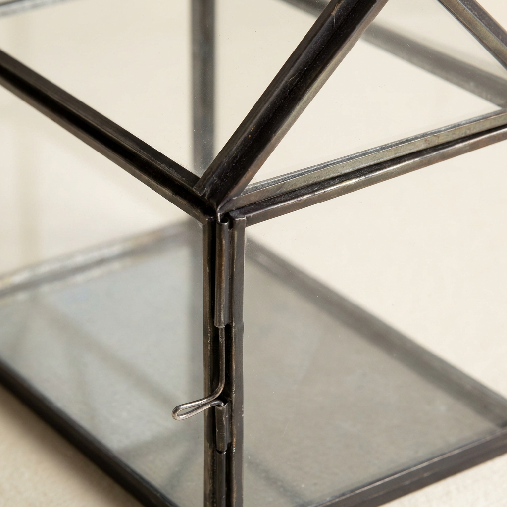 Close-up of the black iron frame, glass roof panels, and side door latch of the miniature greenhouse terrarium.