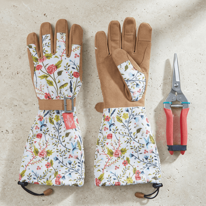 Overhead detail of Garden of Paradise floral gardening gloves with tan palms and red-handled pruning shears on a light stone surface.