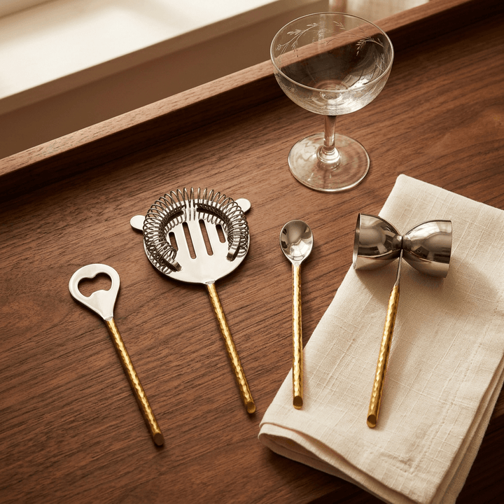 Silver-and-gold bar tools styled on a walnut console beside cream linen and vintage cocktail glassware.