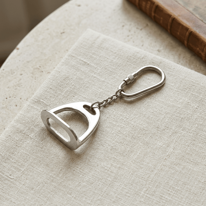 Polished silver-tone stirrup charm keychain with screw-lock ring displayed on cream linen over pale stone.