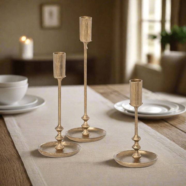 Three antique gold taper candle holders arranged on a linen runner on an oak dining table with white dinnerware.