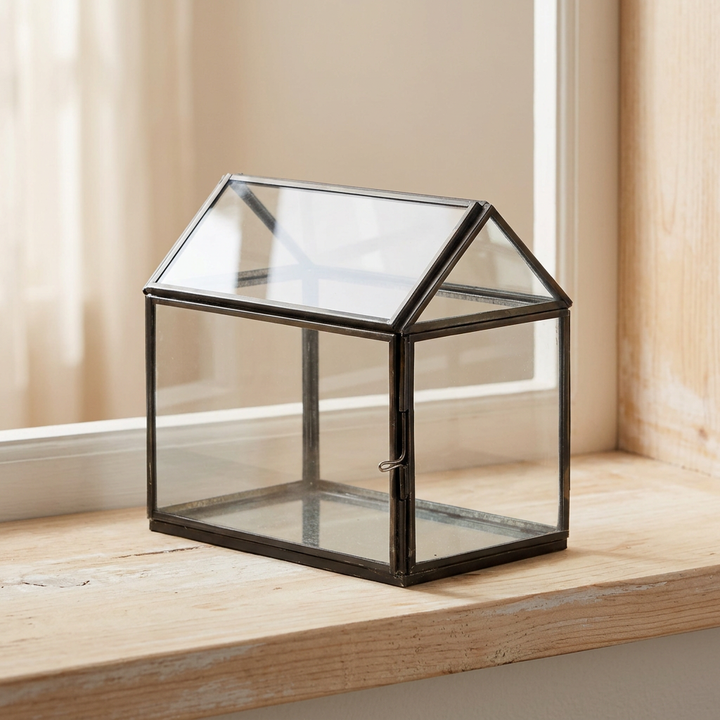 Small empty black-framed glass greenhouse terrarium styled on a pale wood windowsill in soft daylight.