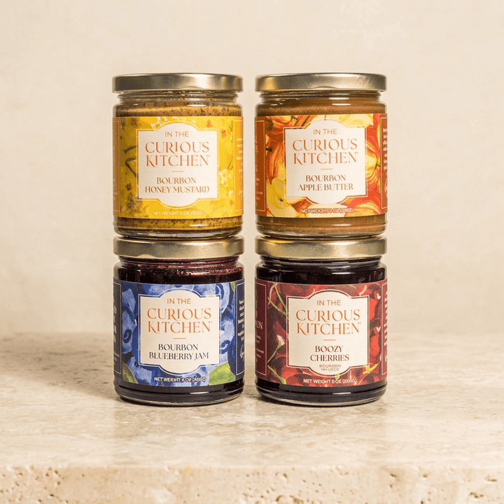 Four gold-lidded jars of bourbon honey mustard, apple butter, blueberry jam, and Boozy Cherries on ivory stone.