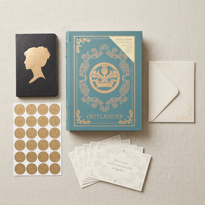 Overhead arrangement of Outlander keepsake book box, black journal, cream notecards, envelopes, and gold stickers on linen.
