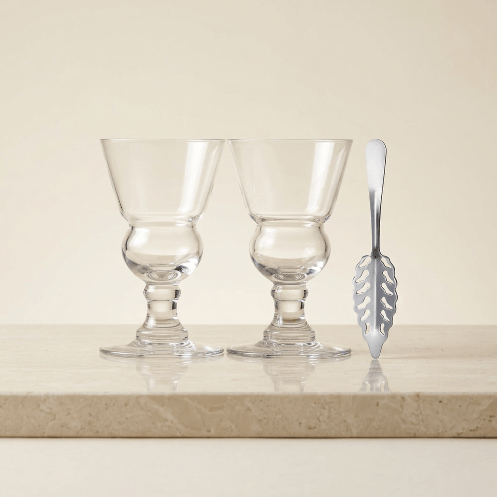 Two empty clear stemmed absinthe glasses with rounded reservoirs and a silver slotted spoon on ivory stone.