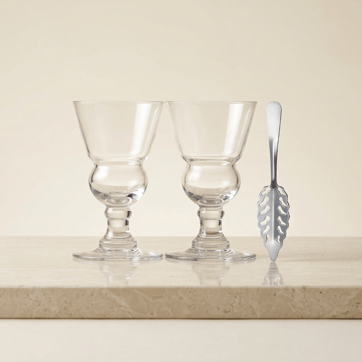 Two empty clear stemmed absinthe glasses with rounded reservoirs and a silver slotted spoon on ivory stone.