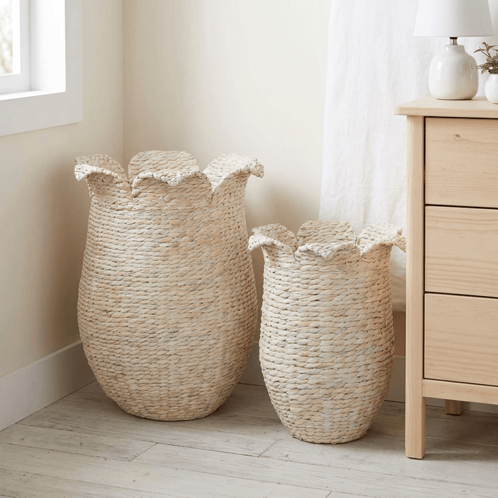 Two petal-rim whitewashed woven storage baskets beside a wooden dresser in a softly lit bedroom.