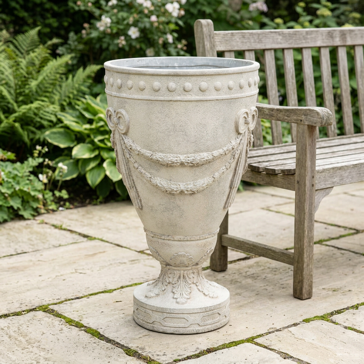 Florentine Floral Urn Planter in a weathered ivory finish on a stone patio beside a wooden garden bench.