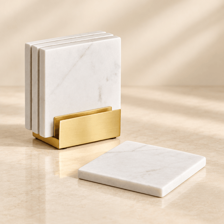 White square marble coasters with a gold metal holder on a warm ivory studio backdrop.