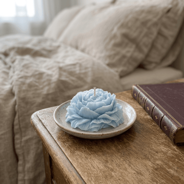 Baby-blue peony-shaped soy wax candle on a ceramic dish beside linen bedding and a book.