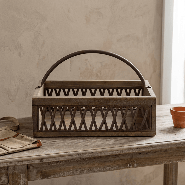 Bramley Garden Caddy, a weathered fir wood and iron lattice garden caddy with an arched iron handle, on a stone garden table beside small potted herbs