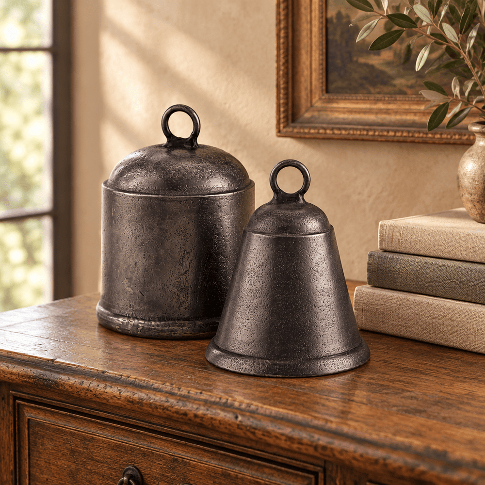 Two antique-pewter bells with circular loop tops styled on a wooden console beside linen-bound books.