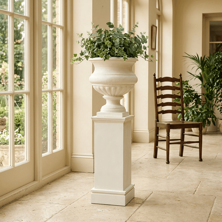 Montclair Iron Urn with Pedestal in Soft White, styled with greenery beside tall sunroom windows.