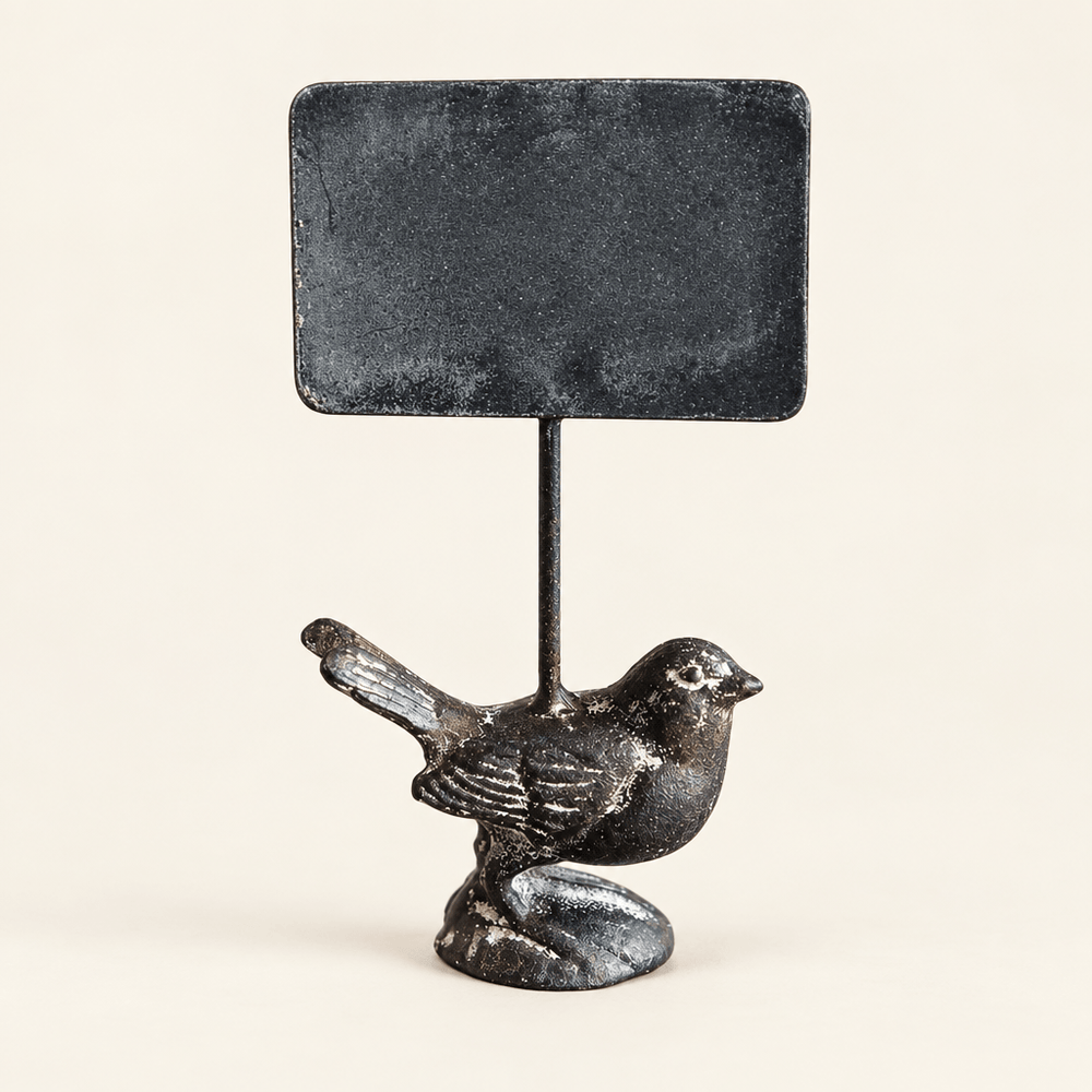 Cast iron bird supporting a blank rectangular chalkboard on an ivory studio backdrop.