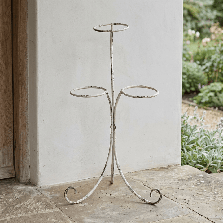 Scrolled iron three-pot plant stand with distressed ivory finish on a stone porch beside a wooden doorway.