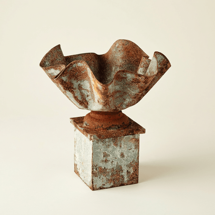 Galvanized iron pedestal planter with a folded rim and rust-brown patina on an ivory studio background.