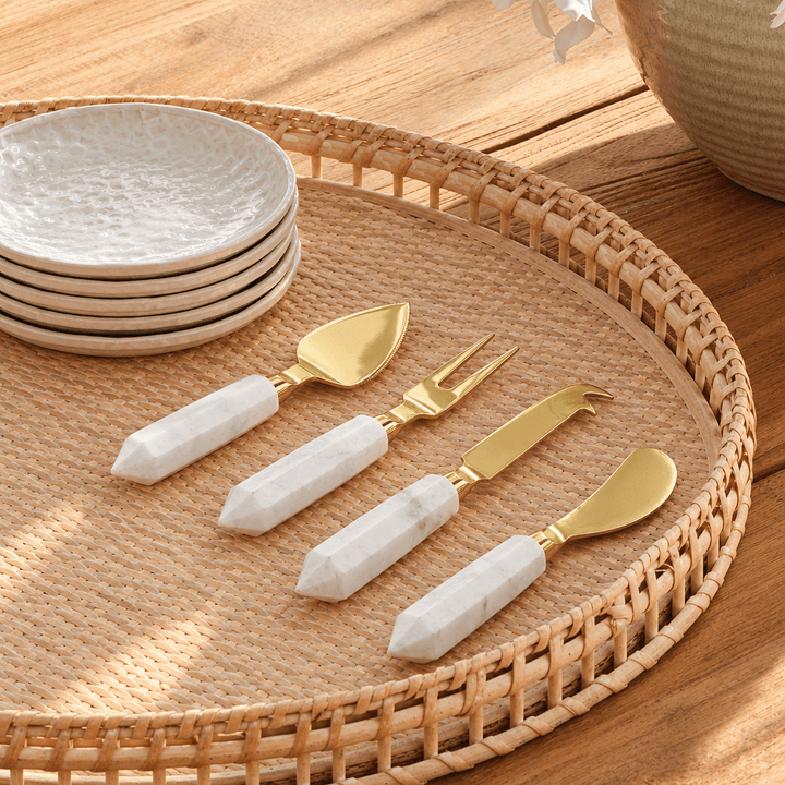 Four marble-handled gold cheese utensils arranged on a woven tray beside ivory ceramic plates